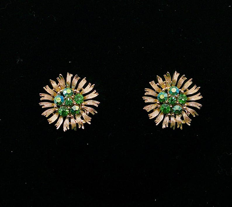 May include: A pair of gold-toned, flower-shaped earrings. Each earring features a cluster of green gemstones at the center, surrounded by radiating, textured gold petals. The earrings are set against a black background.