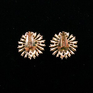 May include: Two gold-toned clip-on earrings with a sunburst design. The earrings have a detailed, textured appearance with a clip-on mechanism on the back. The earrings are set against a black background.