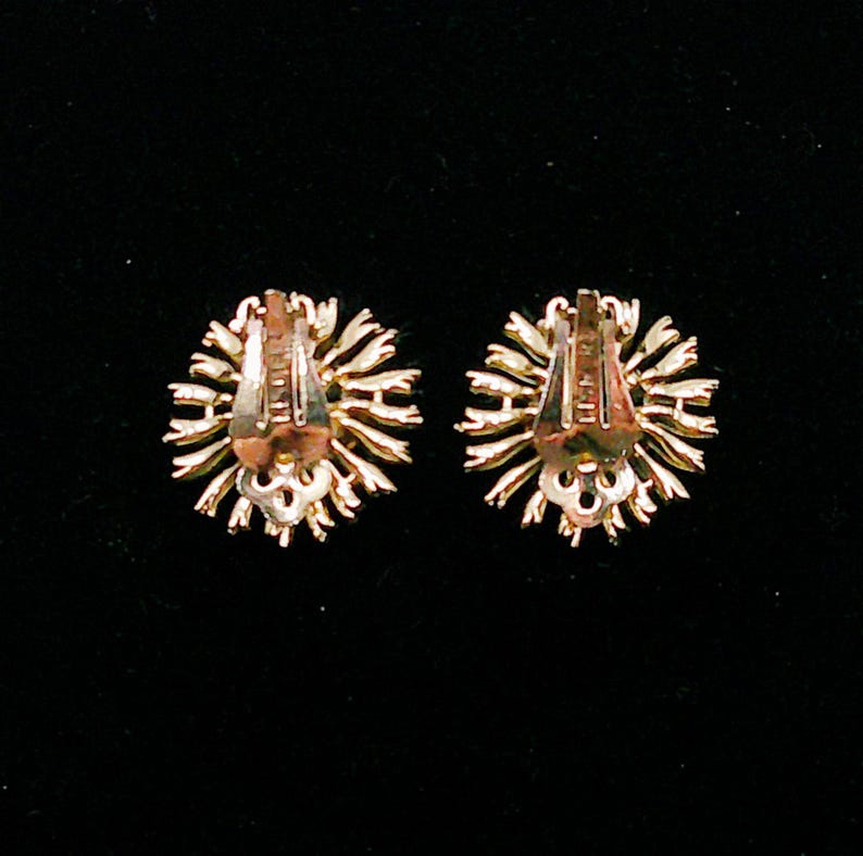 May include: Two gold-toned clip-on earrings with a sunburst design. The earrings have a detailed, textured appearance with a clip-on mechanism on the back. The earrings are set against a black background.