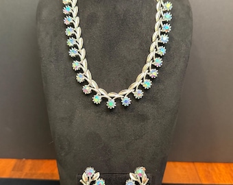 May include: Silver-toned necklace and matching earrings set. The necklace and earrings feature a floral design with iridescent, colorful stones. The jewelry is displayed on a black mannequin, showcasing the intricate details and design.