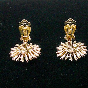May include: A pair of gold-toned clip-on earrings. Each earring features a sunburst design with radiating lines and a clip mechanism. The earrings are set against a dark background.