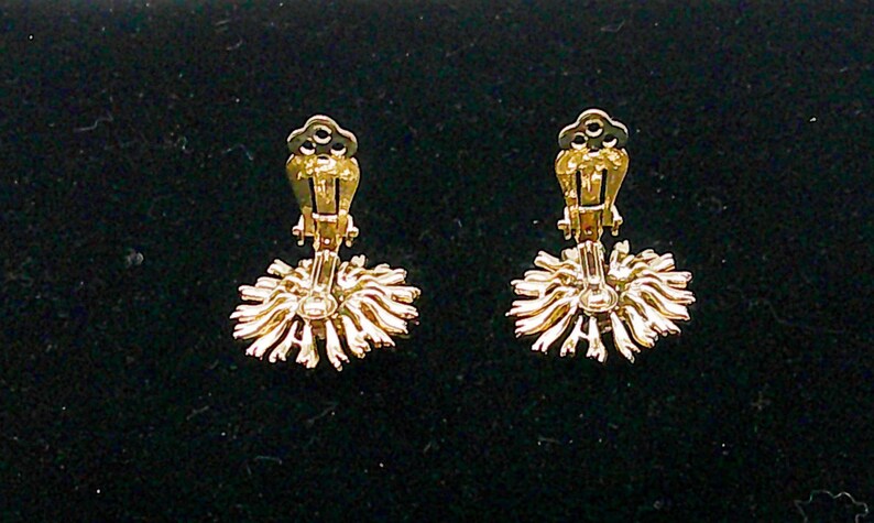 May include: A pair of gold-toned clip-on earrings. Each earring features a sunburst design with radiating lines and a clip mechanism. The earrings are set against a dark background.