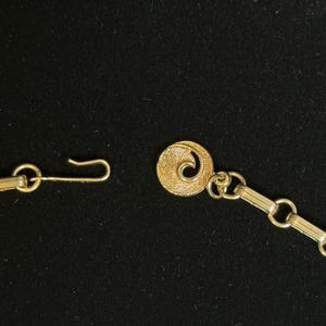 May include: Close-up of a gold-toned necklace clasp and chain against a black background. The clasp features a hook and a decorative circular element with a spiral design. The chain is composed of small, rectangular links.