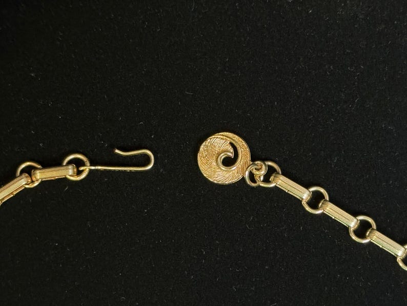 May include: Close-up of a gold-toned necklace clasp and chain against a black background. The clasp features a hook and a decorative circular element with a spiral design. The chain is composed of small, rectangular links.