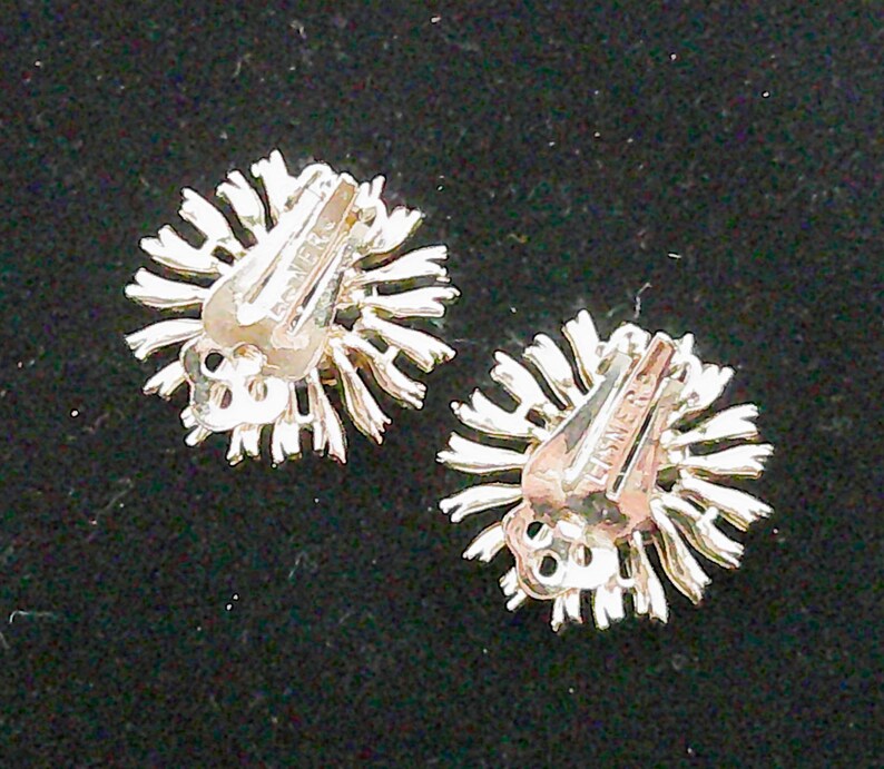 May include: A pair of silver-toned clip-on earrings. Each earring features a sunburst design with radiating lines. The clip mechanism is visible, with the word "EISNER" stamped on the clip.