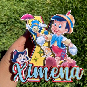 May include: A layered 3D decorative letter K featuring Pinocchio, Jiminy Cricket with a pink umbrella, Figaro the cat, and Cleo the goldfish. The name Ximena is written in blue glittery script across the front, accented with gold star cutouts.