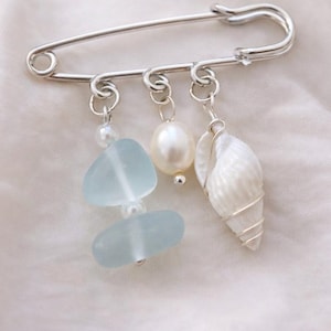 May include: A silver safety pin with three dangling charms. The charms include two light blue sea glass pieces, a white pearl, and a white seashell. The pin is displayed on a white, textured fabric.