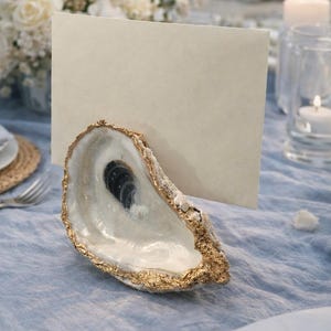 May include: An oyster shell with gold trim holds a blank cream-colored card, set on a light blue tablecloth. The shell is open, revealing a pearlescent interior. White flowers and candles are in the background, suggesting a beach-themed event.