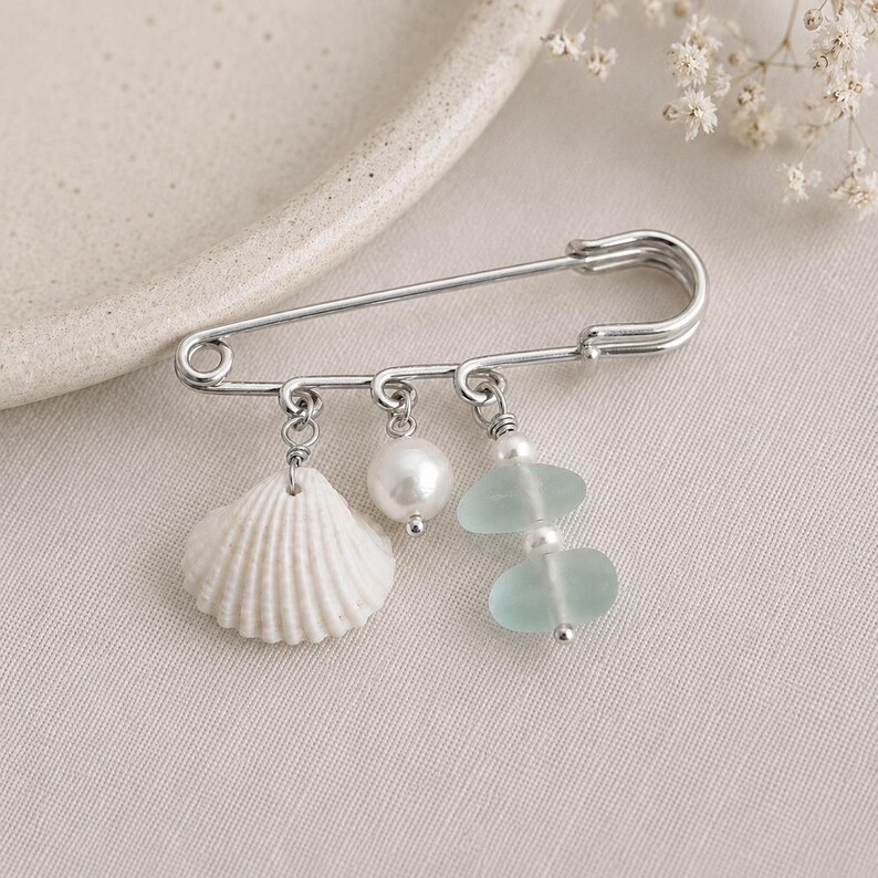 May include: A silver-toned safety pin brooch featuring a white seashell, a faux pearl, and two light blue sea glass pieces. The brooch is a decorative accessory, perfect for adding a touch of coastal charm to clothing or accessories.