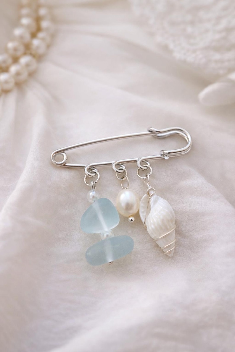 May include: A silver safety pin with three dangling charms. The charms include two light blue sea glass pieces, a white pearl, and a white seashell. The pin is displayed on a white, textured fabric.