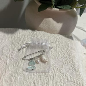 May include: A silver safety pin with dangling charms, including sea glass, a pearl, and a small seashell, presented in a sheer white organza bag. The pin is displayed on a white, textured surface, suggesting a craft or jewelry item.