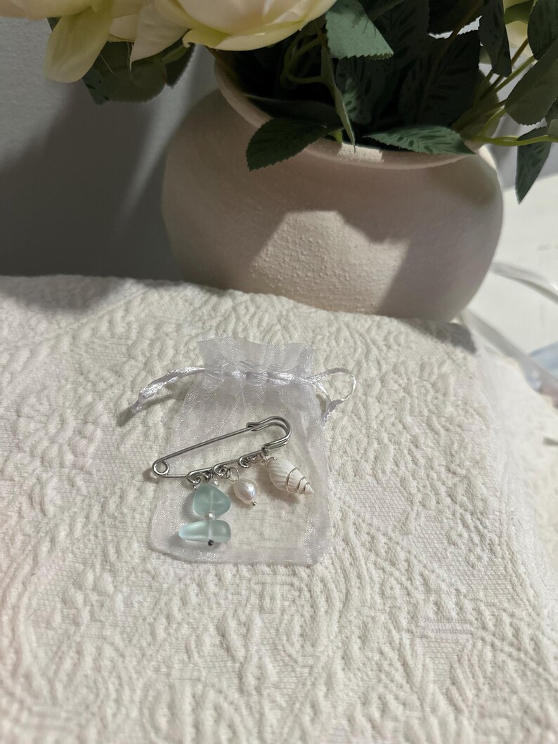 May include: A silver safety pin with dangling charms, including sea glass, a pearl, and a small seashell, presented in a sheer white organza bag. The pin is displayed on a white, textured surface, suggesting a craft or jewelry item.