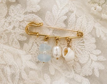 May include: A gold-colored safety pin with three dangling charms: light blue beads, a pearl, and a small seashell. The pin is displayed on a white lace fabric, with delicate floral details.