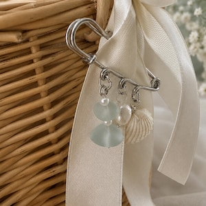 May include: A silver safety pin with dangling charms, including pale blue sea glass, pearls, and a small seashell, is attached to a cream-colored ribbon. The ribbon is tied to a woven wicker basket, suggesting a beach or nautical theme.