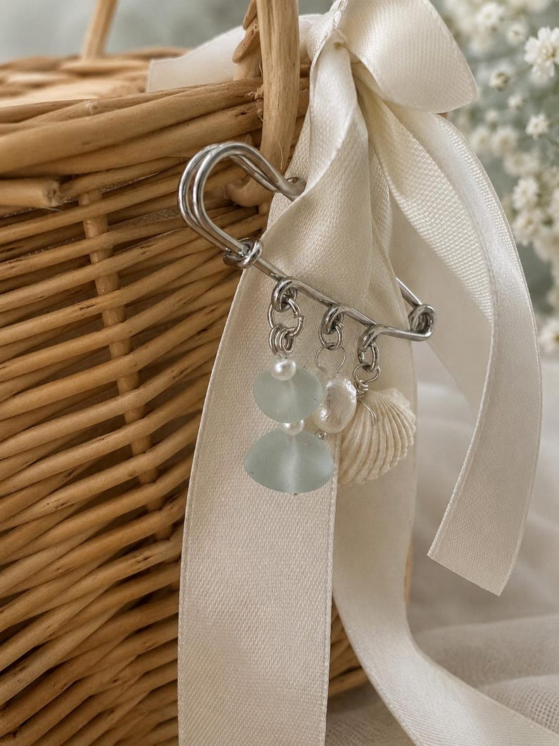 May include: A silver safety pin with dangling charms, including pale blue sea glass, pearls, and a small seashell, is attached to a cream-colored ribbon. The ribbon is tied to a woven wicker basket, suggesting a beach or nautical theme.