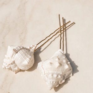 May include: Two white seashell hair pins with gold-colored metal stems. The seashells have a textured surface and are arranged on a light-colored background. The pins are designed for hair styling.