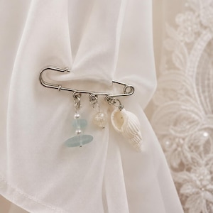 May include: A silver safety pin with three dangling charms. The charms include a seashell, a clear heart-shaped bead, and a stack of blue-green beads. The pin is attached to white fabric, suggesting a decorative or accessory item.