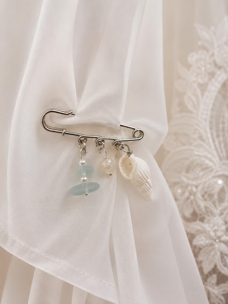 May include: A silver safety pin with three dangling charms. The charms include a seashell, a clear heart-shaped bead, and a stack of blue-green beads. The pin is attached to white fabric, suggesting a decorative or accessory item.