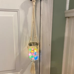 May include: A hanging mason jar with colorful fairy lights inside. The jar is suspended by a cream-colored macrame plant hanger with a tassel. The jar is hanging from a black door handle.