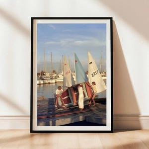 May include: A framed wall art print depicting three individuals on a wooden dock maneuvering a small sailboat with a light blue sail. In the background, several other sailboats are moored in a harbor under a clear sky with soft daylight.