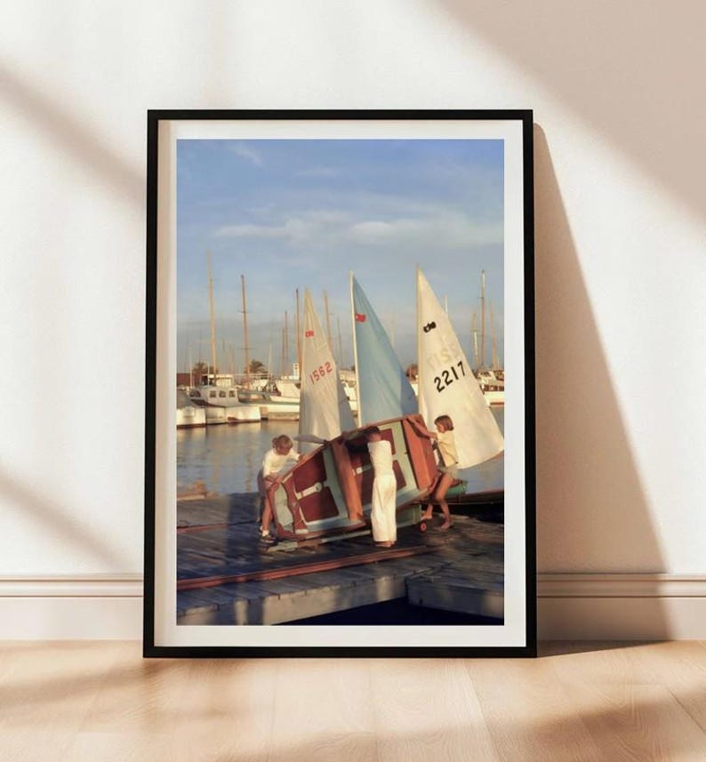 May include: A framed wall art print depicting three individuals on a wooden dock maneuvering a small sailboat with a light blue sail. In the background, several other sailboats are moored in a harbor under a clear sky with soft daylight.