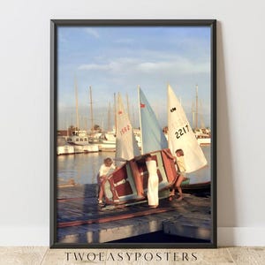 May include: A framed wall art print depicting two individuals in white clothing moving a small boat on a wooden dock. In the background, several sailboats with white and light blue sails are moored in a harbor under a clear sky. The text TWOEASYPOSTERS is visible at the bottom of the frame.