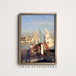 May include: A framed vertical poster featuring a vintage photograph of three individuals on a wooden dock preparing a small sailboat. In the background, several sailboats with white and blue sails are moored in a harbor under a clear sky.