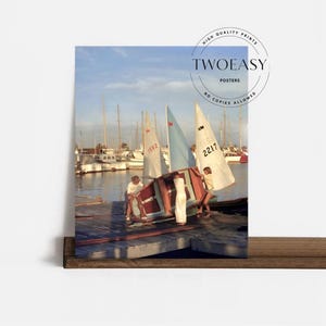 May include: A photographic print titled TWOEASY POSTERS featuring two individuals in white clothing maneuvering a small sailboat with a light blue and white sail on a wooden dock, with numerous other sailboats moored in the background water.