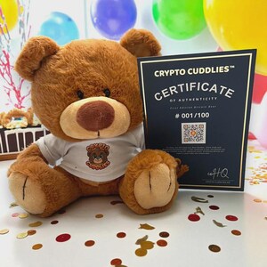 May include: A brown teddy bear wearing a white t-shirt with a logo that says "Crypto Cuddlies". The bear is next to a certificate of authenticity with the text "Crypto Cuddlies" and "First Edition Bitcoin Bear". The certificate is numbered 001/100.