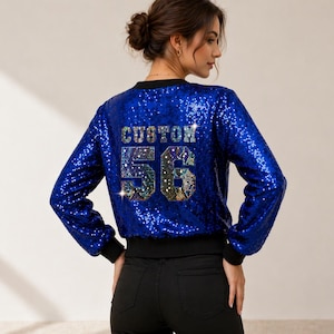 Pode incluir: Casaco bomber azul com lantejoulas, mangas compridas e acabamento preto. A parte de trás do casaco apresenta a palavra "CUSTOM" acima do número "56" num design multicolorido e brilhante. O casaco é uma peça vibrante e apelativa.