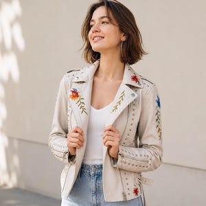 May include: Cream-colored leather jacket with floral embroidery in red, orange, and blue. The jacket features silver studs, a notched collar, and a zipper closure. The model is wearing a white top and blue jeans.