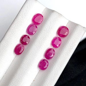 Könnte beinhalten: Sieben oval geschliffene Rubine mit einem leuchtenden, tief rosa-roten Farbton und facettierten Oberflächen. Die Edelsteine sind in zwei vertikalen Reihen auf einem weißen Präsentationsständer angeordnet und zeigen ihre Klarheit und Form.