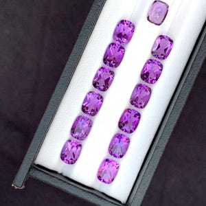 Könnte beinhalten: Eine Sammlung von elf kissenförmig geschliffenen violetten Edelsteinen, die in zwei Reihen in einer schwarzen Präsentationsbox mit weißem Samteinsatz angeordnet sind. Jeder Edelstein hat eine facettierte Oberfläche, die das Licht reflektiert.