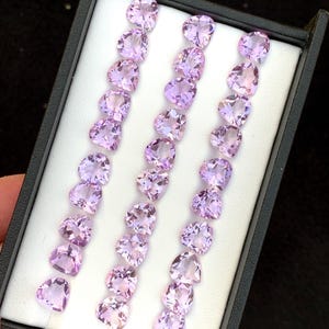 Könnte beinhalten: Eine Sammlung von hellvioletten, herzförmigen facettierten Edelsteinen, die in drei vertikalen Reihen in einer schwarzen Präsentationsbox mit weißem Innenfutter angeordnet sind. Die Steine sind durchscheinend und haben eine polierte Oberfläche.