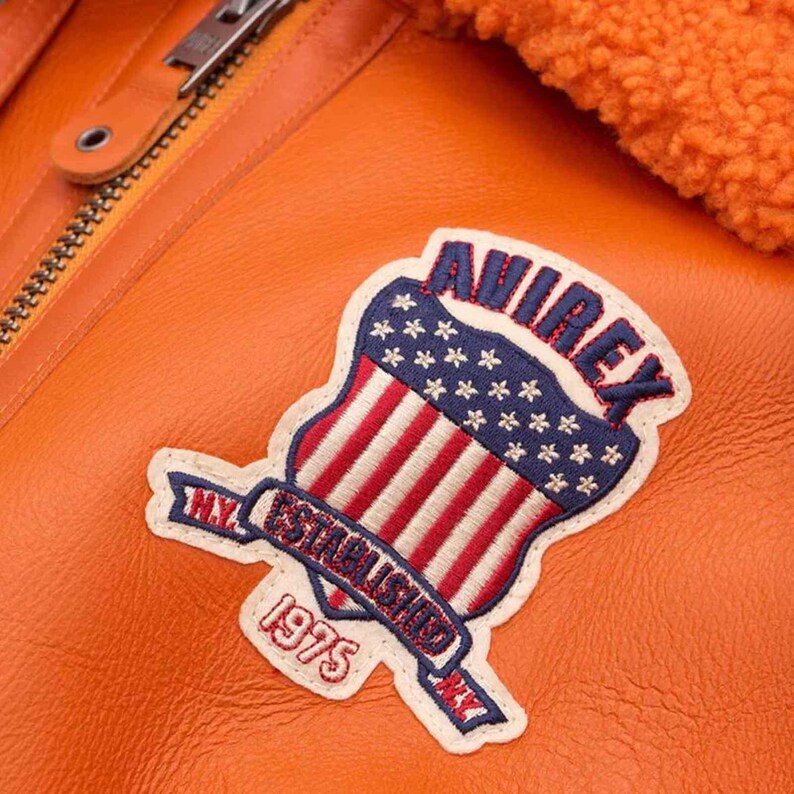 May include: Close-up of an orange leather jacket with a patch. The patch features a shield design with red and white stripes, blue stars, and the word "AVIREX". The patch also includes the text "N.Y. ESTABLISHED 1975".