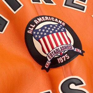 May include: Close-up of an orange leather jacket with patches. The main patch features a shield design with the text "ALL AMERICANS ESTABLISHED 1975 NY" and a red, white, and blue color scheme. Other patches with black and white text are also visible.