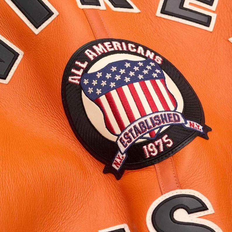 May include: Close-up of an orange leather jacket with patches. The main patch features a shield design with the text "ALL AMERICANS ESTABLISHED 1975 NY" and a red, white, and blue color scheme. Other patches with black and white text are also visible.