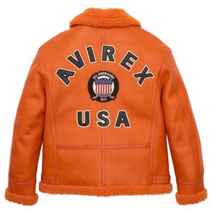 May include: Orange aviator jacket with a shearling collar and lining. The back features the word "AVIREX" above a circular patch with the American flag and the word "USA" below. The jacket has a classic design with a warm, inviting aesthetic.