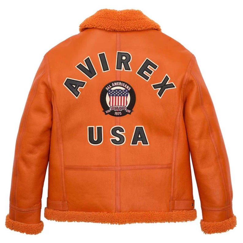 May include: Orange aviator jacket with a shearling collar and lining. The back features the word "AVIREX" above a circular patch with the American flag and the word "USA" below. The jacket has a classic design with a warm, inviting aesthetic.