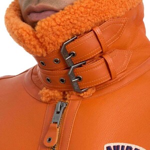 May include: Close-up of an orange leather jacket with a shearling collar. The jacket features a double-buckle closure at the neck and a zipper down the front. The collar and lining are a contrasting orange color.