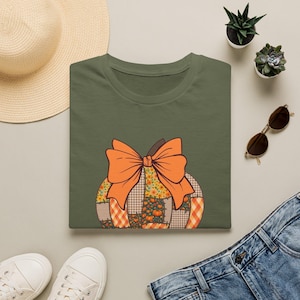 Puede incluir: Una camiseta verde oliva con un gráfico de una calabaza hecha de varios patrones de patchwork, incluyendo cuadros, vichy y estampados florales, rematada con un gran lazo naranja sólido. El diseño está centrado en la parte frontal de la prenda.