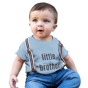 May include: A baby wearing a light blue t-shirt with the words "little brother" printed on the front. The shirt is paired with blue denim jeans and suspenders with brown leather and striped detailing.