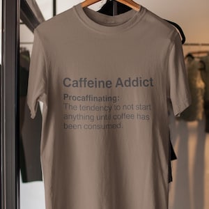 Puede incluir: Una camiseta color arena con el texto "Caffeine Addict" y la definición de "Procaffinating" en letras grises. La camiseta está colgada en una percha de madera.