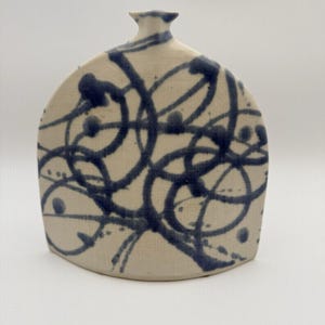 May include: A flat, rounded ceramic vase with a cream-colored base and a dark blue hand-painted pattern of overlapping circular lines and dots. The vessel features a narrow, short neck at the top and a wide, curved body with a flat base for stability.