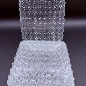 May include: A set of two square clear glass plates featuring a repeating pressed starburst and hobnail pattern across the surface. Each plate has a scalloped edge with a decorative border, creating a textured, reflective appearance throughout.