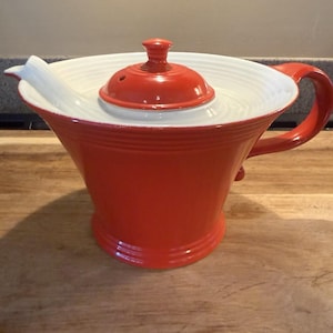May include: A bright orange ceramic teapot featuring a wide, white interior basin with concentric ring detailing. The teapot includes a matching orange lid with a small knob handle and a curved orange side handle, showcasing a glossy finish.