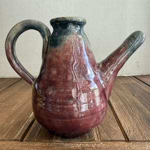 Puede incluir: Una jarra de cerámica con acabado brillante, cuerpo redondeado y un pico largo e inclinado. El esmalte muestra una mezcla de tonos burdeos profundo y marrón rojizo, que transicionan a un verde oscuro y apagado en el cuello y el asa.