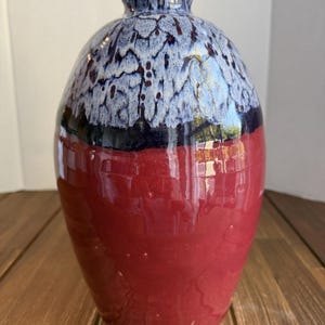 May include: A ceramic vase with a glossy finish, featuring a deep red base that transitions into a dark navy band, topped with a textured white and blue drip glaze pattern. The vessel has a rounded body and a narrow, short neck opening.