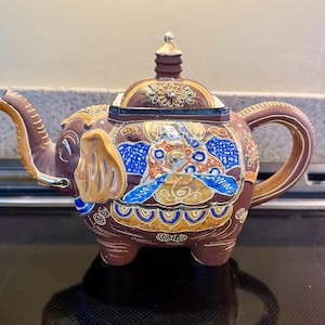 May include: A brown ceramic teapot shaped like an elephant with a matching lid. The body features intricate hand-painted details in blue, orange, gold, and white, including a central figure, floral motifs, and decorative patterns along the base.