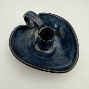 Puede incluir: Un portavelas de cerámica en forma de corazón con un esmalte azul oscuro brillante. La pieza cuenta con un receptáculo cilíndrico central para una vela y un asa curva unida al borde, creando un diseño funcional con un acabado liso y brillante.